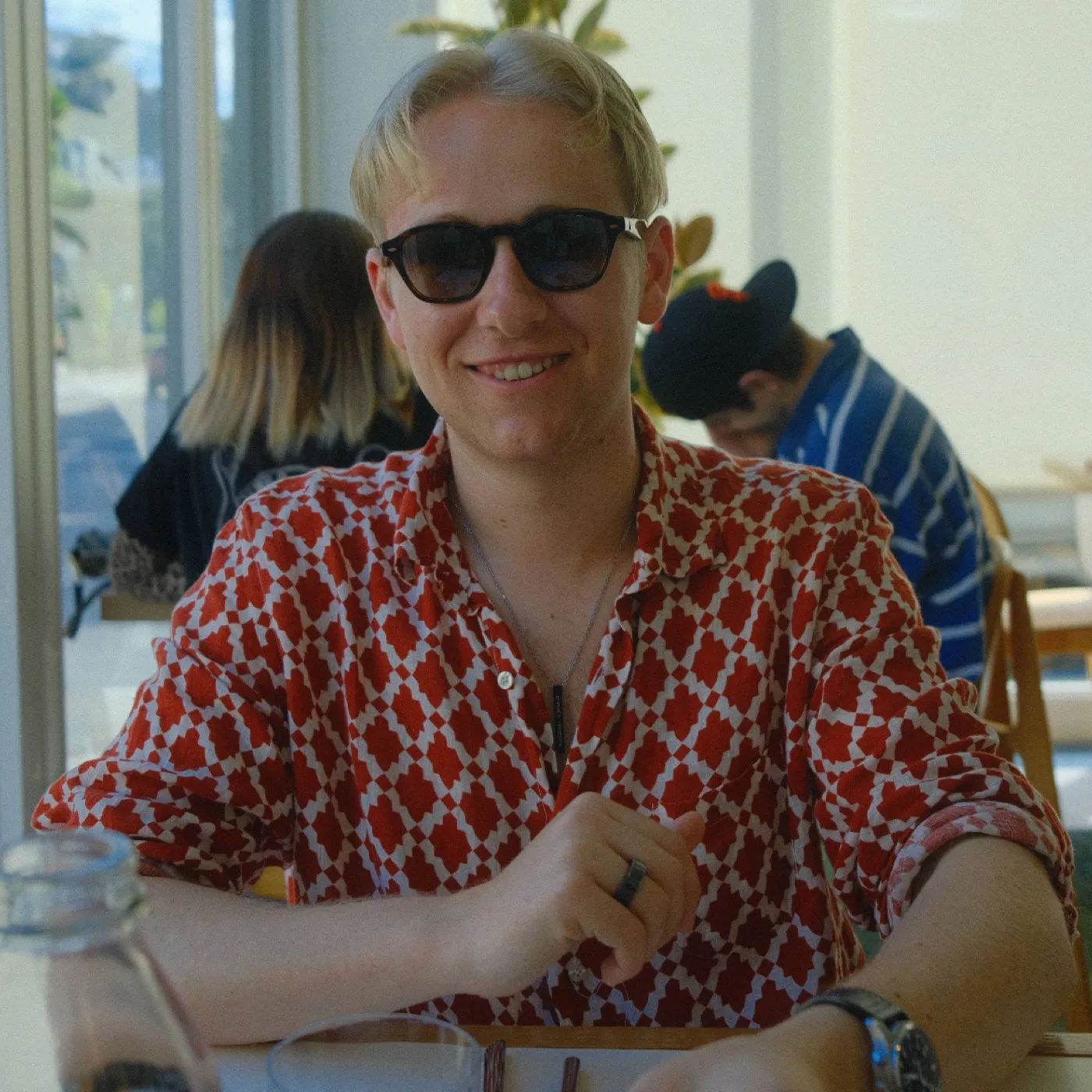 Raffael outdoors in bright daylight, in sunglasses and a patterned shirt, smiling at the camera.